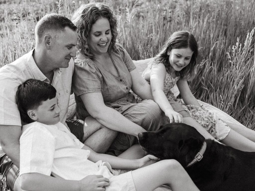 Family of four sitting on a blanket in a field with their dog at a family photo session. 